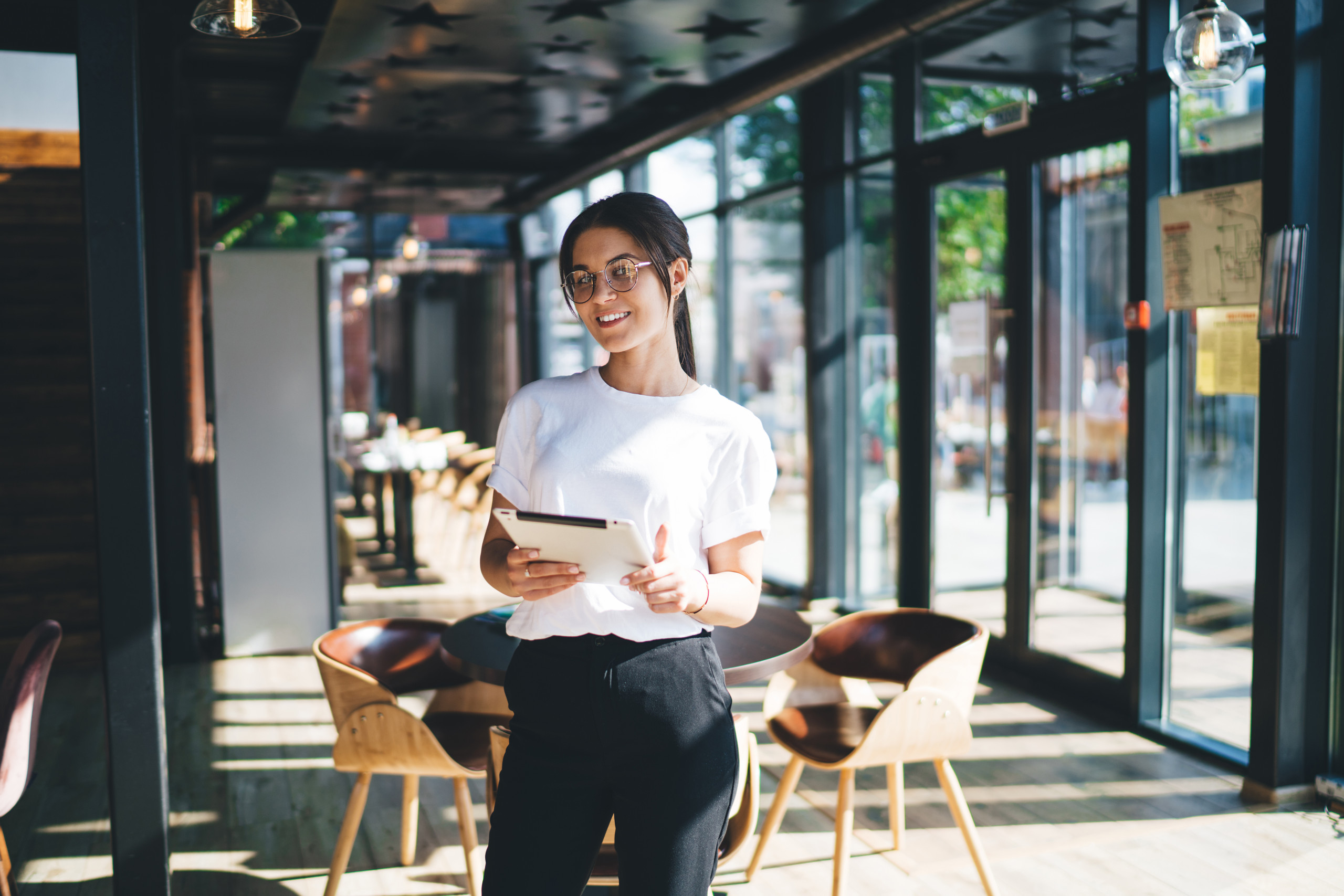 Jeune hôtesse de restaurant souriante, vêtue d’une tenue décontractée et portant des lunettes, regardant l’objectif tout en se tenant debout devant un intérieur chaleureux flouté, tenant une tablette et souriant.